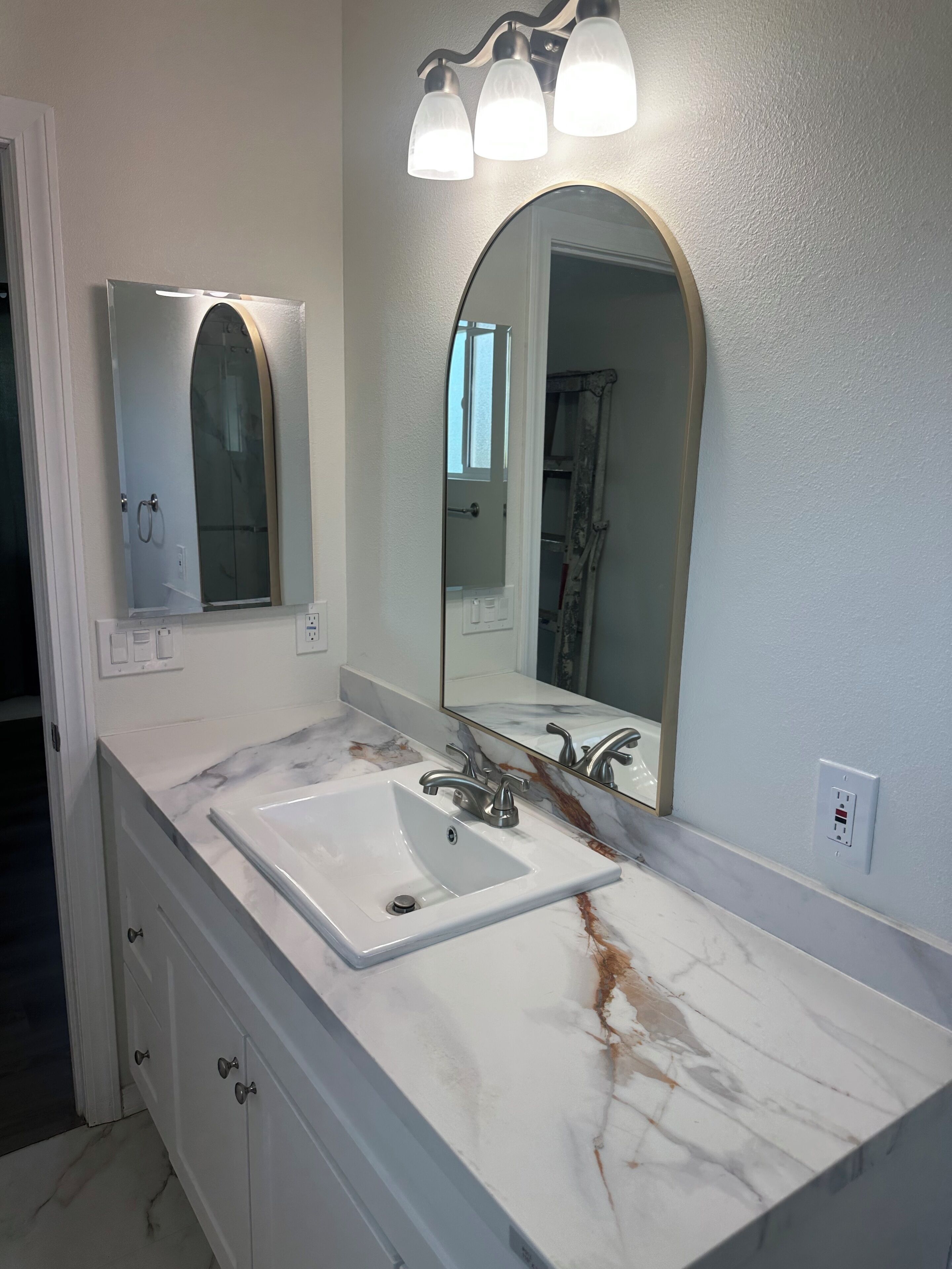 Une salle de bain avec un lavabo en marbre blanc, un grand miroir surmonté d’un arc et un luminaire fixé au mur.