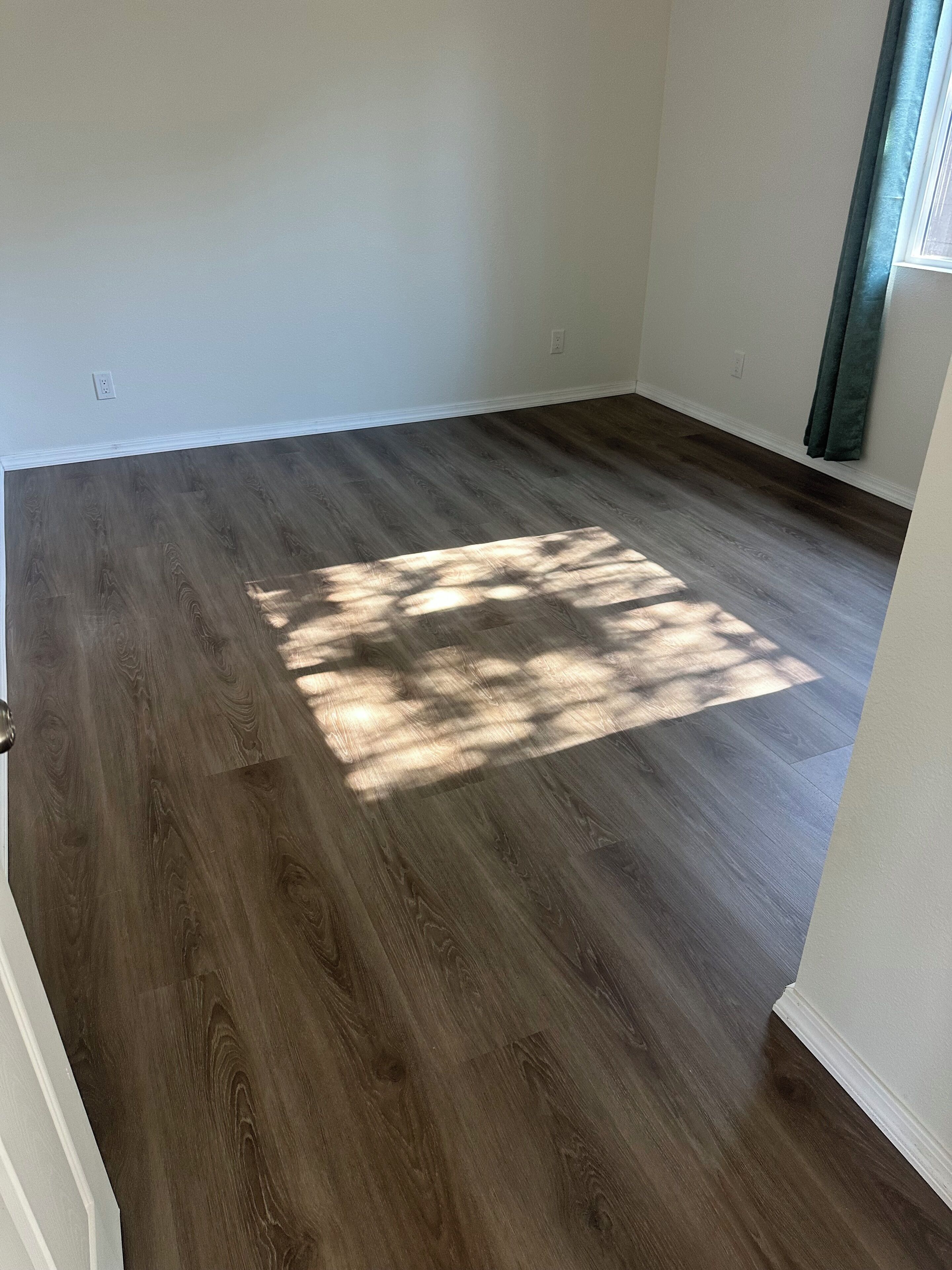 Una habitación con piso de madera oscura, paredes blancas y una ventana con cortina.