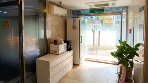 Zona de recepción con puerta de vidrio, mostrador de recepción, planta en maceta y lámpara en el techo.