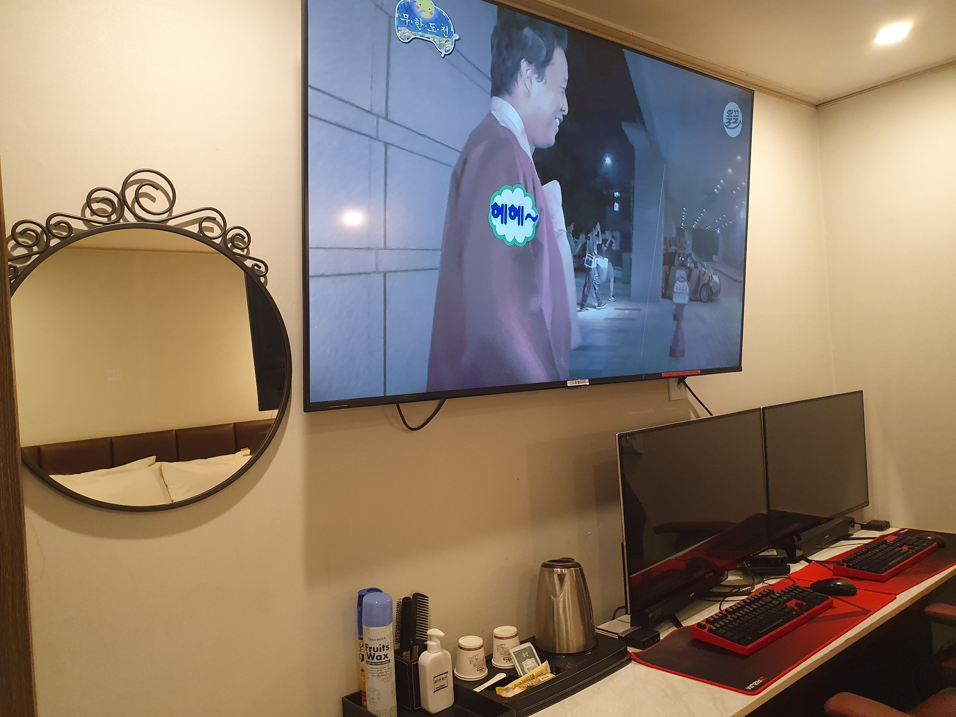 Habitación de hotel con una amplia televisión de pantalla plana montada en la pared, una cama con cabecera, un escritorio con computadora, teclado, ratón y varios objetos personales.