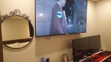 Habitación de hotel con una amplia televisión de pantalla plana montada en la pared, una cama con cabecera, un escritorio con computadora, teclado, ratón y varios objetos personales.