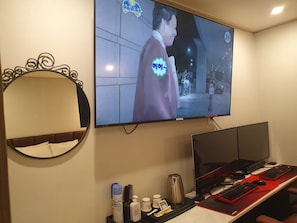Habitación de hotel con una amplia televisión de pantalla plana montada en la pared, una cama con cabecera, un escritorio con computadora, teclado, ratón y varios objetos personales.