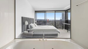 Un dormitorio moderno con una cama grande, una mesita de noche, un tocador y un baño con bañera, además de una ventana con vista a la ciudad.