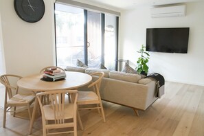 A living room with a round wooden table, four chairs, a beige sofa, a TV mounted on the wall, and a large window with blinds. - Charming 2BDR Apartment in Richmond close to MCG (Richmond)