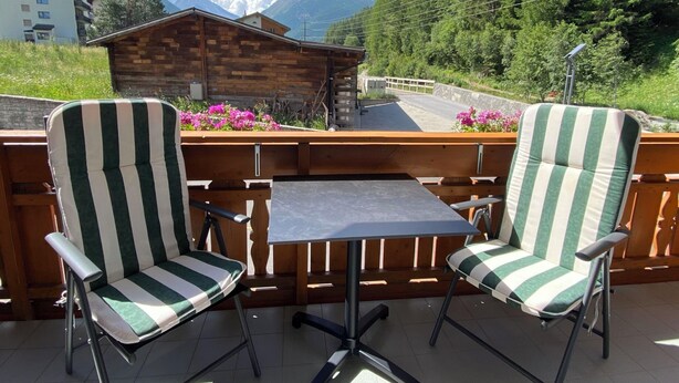 A balcony with two striped chairs and a table, overlooking a mountain landscape and a wooden building. - Haus Saturn Studio STUDIO by Interhome (Saas-Grund)