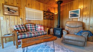 A wooden cabin interior with a plaid sofa, a leather armchair, a wood-burning stove, and a framed duck painting.