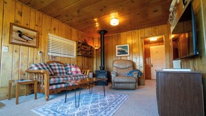 A wooden cabin interior with a plaid sofa, a leather chair, a fireplace, and a wooden cabinet.
