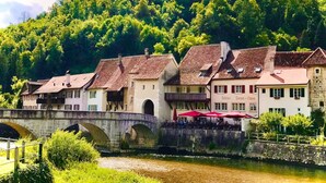 Un hotel a orillas del río con arquitectura tradicional, un puente de piedra y exuberante vegetación al fondo.