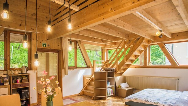 Interior de madera con una cama, un escritorio con un florero y una escalera, con vistas a la vegetación exterior.