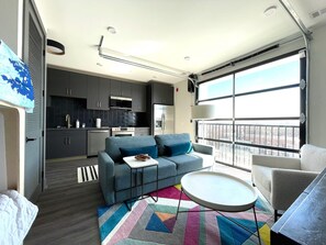Ein kompakter Wohnraum mit Küchenzeile, einer Couch, einem kleinen Tisch und einem großen Fenster mit Blick.