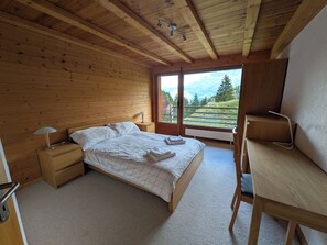Ein Schlafzimmer mit Holzbalkendecke, einem Bett mit weißer Bettwäsche, einem Nachttisch mit Lampe, einem Schreibtisch und Blick auf eine Berglandschaft durch eine Glastür.