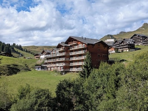 Ein mehrstöckiges Hotel mit Balkonen, gelegen in einer gebirgigen Landschaft mit grünen Hügeln und Bäumen.
