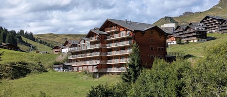 A multi-story wooden building with balconies, set in a mountainous area with green hills and a clear sky.
