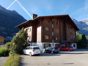 A multi-story building with a sloped roof, parked cars in front, and mountains in the background. - Résid. Mila 5, (Champery), by Interhome (Champery)