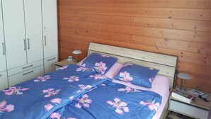Ein Schlafzimmer mit hölzernem Kopfende, einem Bett mit floralem Bettzeug, einem Nachttisch mit Lampe und einem Kleiderschrank.