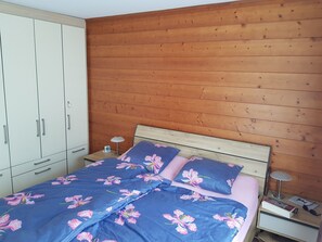 A bedroom with a wooden headboard, a bed with floral bedding, a nightstand with a lamp, and a wardrobe. - Résid. Mila 5, (Champery), by Interhome (Champery)