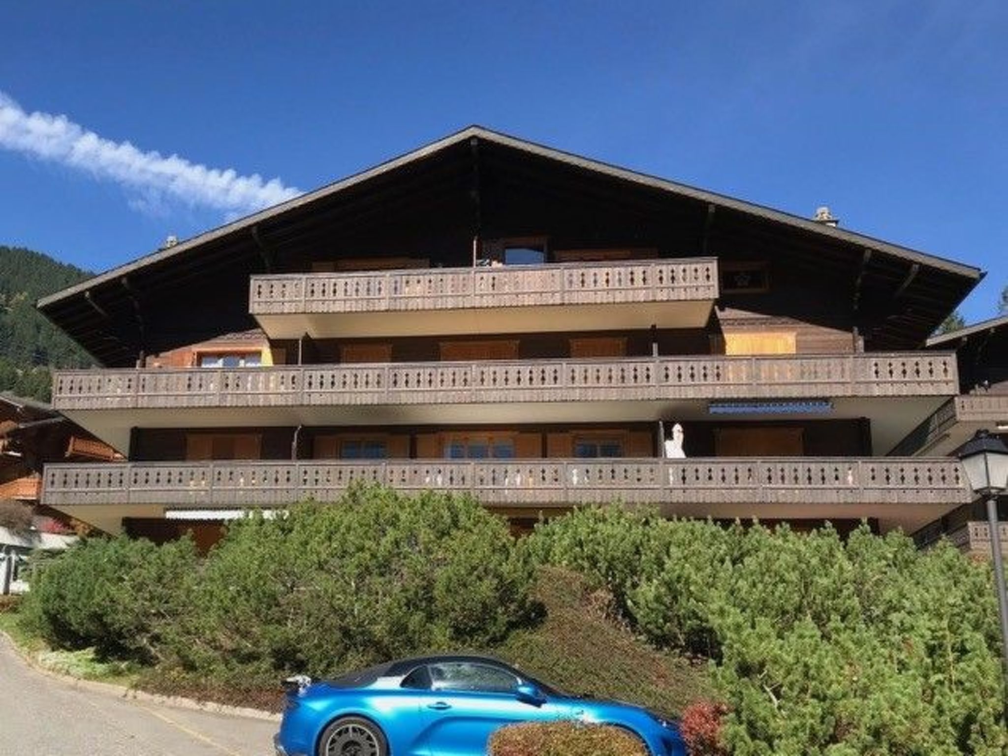 A multi-story building with balconies, a car parked in front, and a mountainous backdrop.