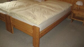 Ein hölzernes Bett mit weißer Matratze und Kissen, ein Nachttisch aus Holz mit einer Lampe sowie ein Heizkörper im Hintergrund.