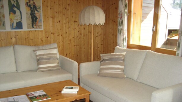 Ein Zimmer mit Holzboden, einem weißen Sofa mit gestreiften Kissen, einem Holztisch mit Büchern und einer Fernbedienung sowie einer weißen Lampe.