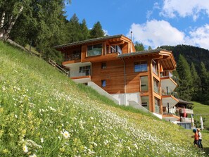 Une maison moderne en bois, à plusieurs niveaux, située sur une colline, avec un jardin en contrebas.
