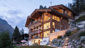 A multi-story wooden building with balconies, a stone wall, and a sign reading 'CHALET NE POMUK.' - Chalet Nepomuk, (Zermatt), 1632.00 by Interhome (Zermatt)