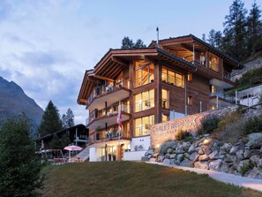 A multi-story wooden building with balconies, a stone wall, and a sign reading 'CHALET NE POMUK.' - Chalet Nepomuk, (Zermatt), 1632.00 by Interhome (Zermatt)