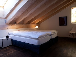 A bedroom with a sloped ceiling, a bed, a bedside table, a lamp, and a chair. - Chalet Altesse, (Zermatt), 1054.01 by Interhome (Zermatt)