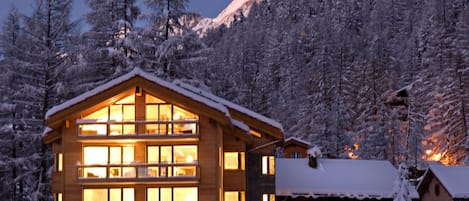 A wooden lodge with multiple windows and a snow-covered roof, surrounded by snow-laden trees and a mountain in the background.