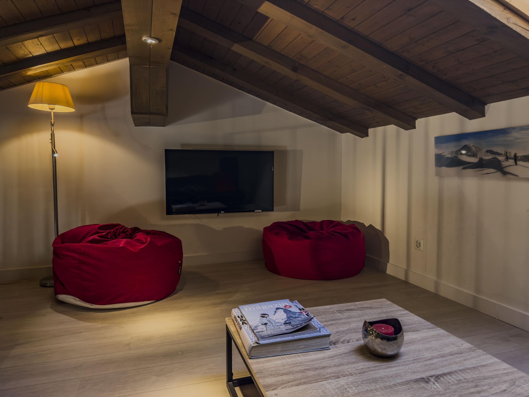 Ein gemütliches Zimmer mit Holzbalken, einem Flachbildfernseher, zwei roten Sitzsäcken, einer Zeitschrift und einer Kerze auf einem Tisch.