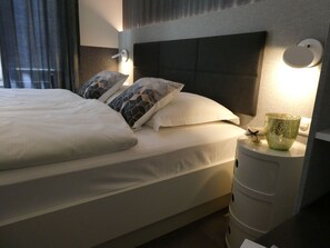 A neatly made bed with white bedding and patterned pillows, a bedside lamp, and a small table with a glass container. - Haus Venus, (Zermatt), by Interhome (Zermatt)