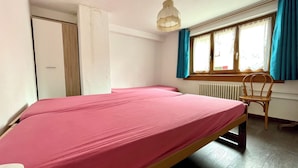 Una cama individual con una colcha rosa en una habitación con ventana, una silla y un radiador.