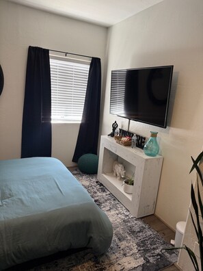 A bedroom with a bed, a TV, a small white console table, a window with curtains, and a green round cushion. - Cozy Music Bedroom Shared Newley Remodeled House. Peaceful, Darksky neighborhood (Clarkdale)