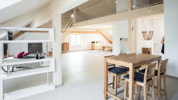 A spacious loft with a high ceiling, exposed wooden beams, a dining area with a wooden table and chairs, and a staircase leading to an upper level. - Appartements - Ferme La Joux-Chaupe, (St-Ursanne), by Interhome (Saint-Ursanne)