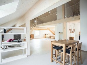 Un loft espacioso con techo alto, vigas de madera a la vista, una zona de comedor con mesa y sillas de madera, y una escalera que lleva a un nivel superior.