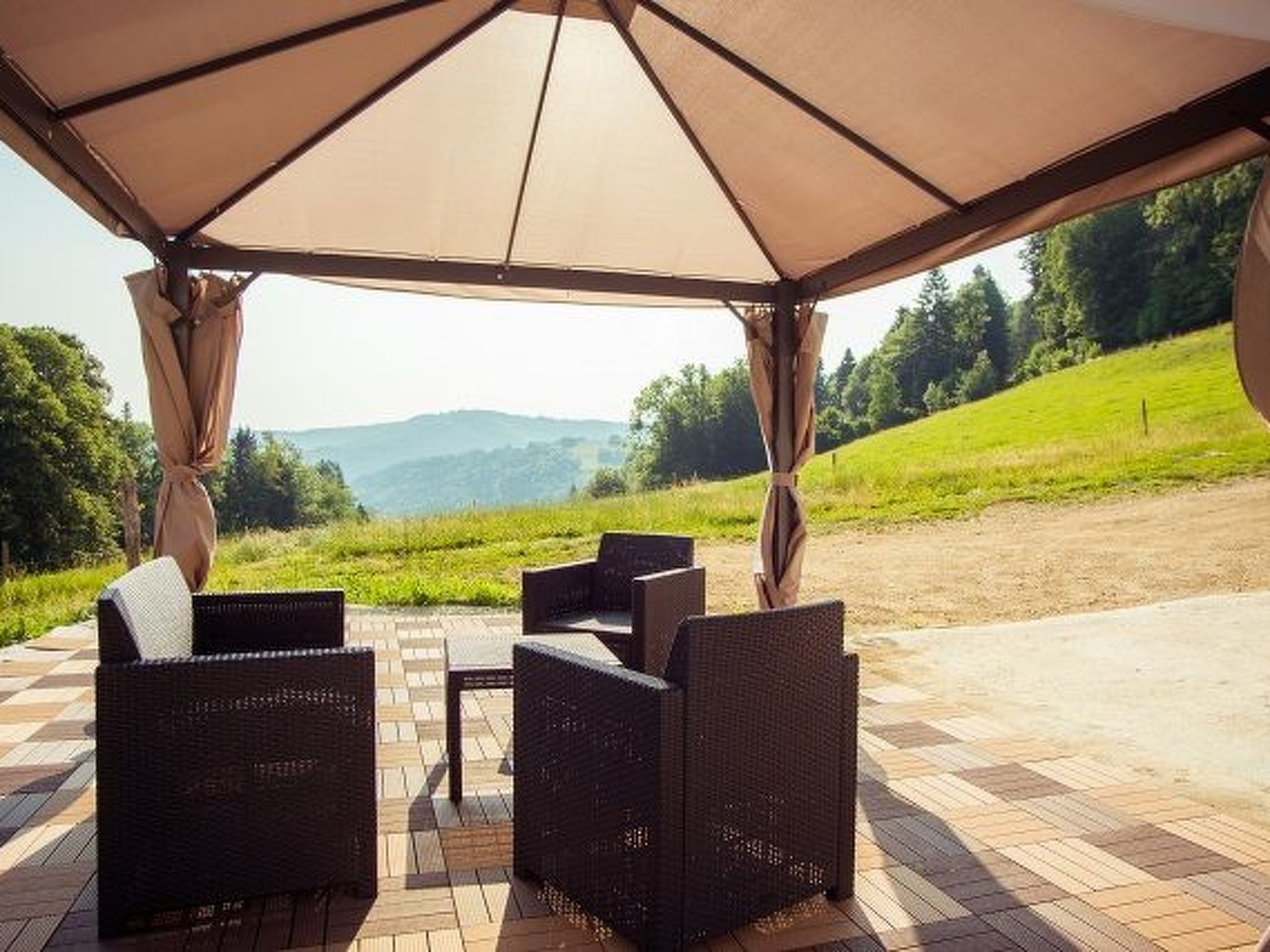 Eine Terrasse im Freien mit überdachter Sitzgruppe, Korbmöbeln und Blick auf eine grüne Landschaft.