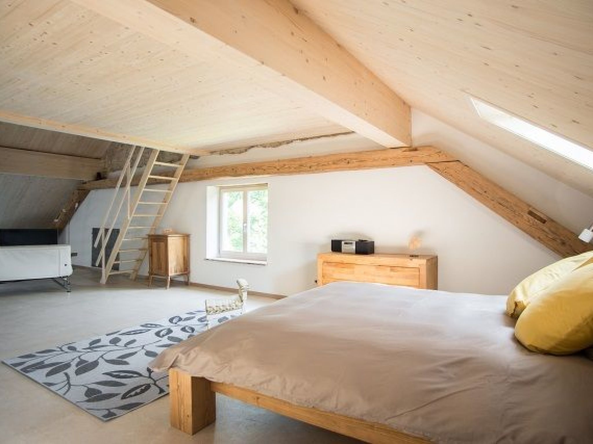 A bedroom with a sloped ceiling, a bed, a bedside table, a nightstand, a window, and a wooden ladder.