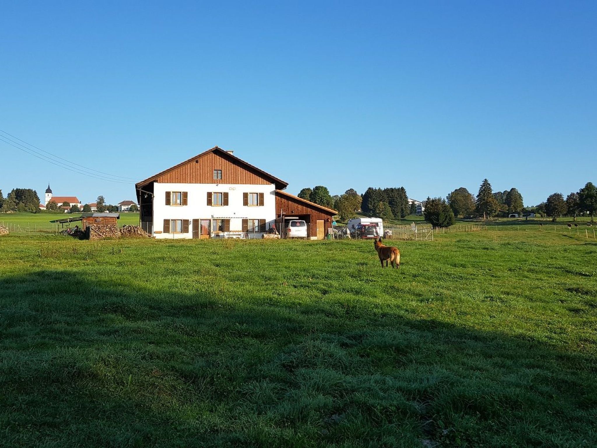 A two-story building with a white facade and brown roof, surrounded by a green field and a few trees.