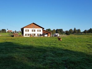 En toetages bygning med en hvid facade og et brunt tag, omgivet af en grøn mark og nogle få træer.