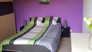A bedroom with a bed, a wall-mounted picture, a white vase with flowers, and a white ceiling light. - Appartement - Sous les Barres, (Montfaucon), by Interhome (Montfaucon)