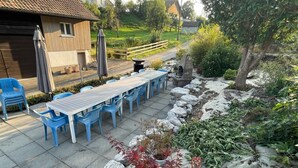 Un patio exterior con una mesa larga y sillas azules, rodeado de rocas y plantas.