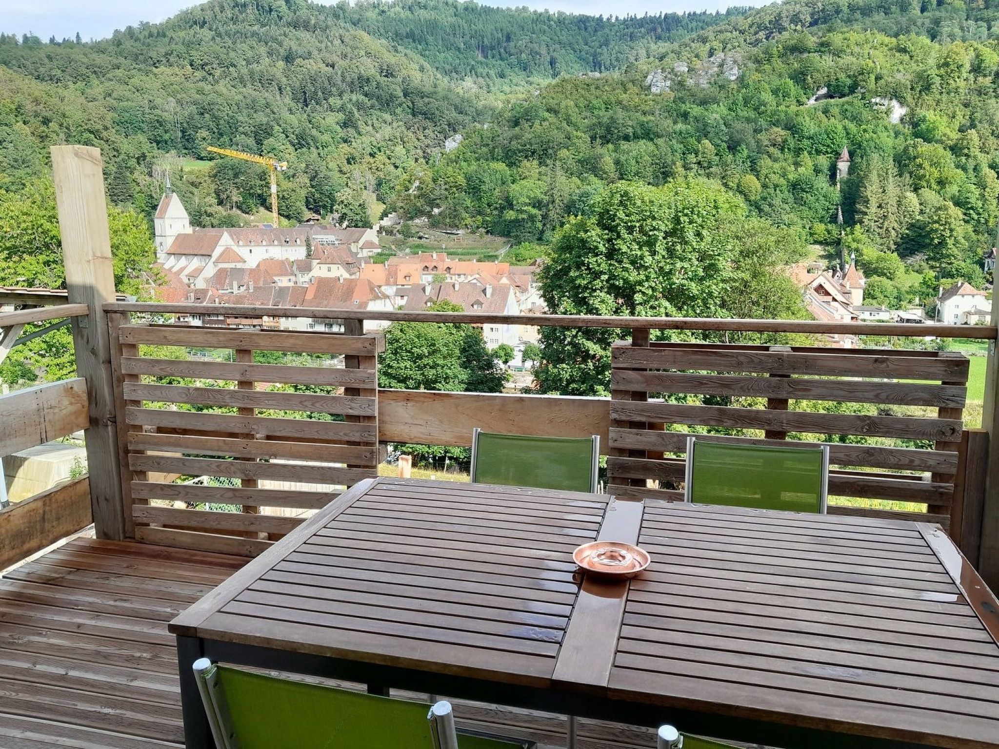 Une terrasse en bois avec une table et des chaises, donnant sur une colline boisée parsemée de bâtiments.