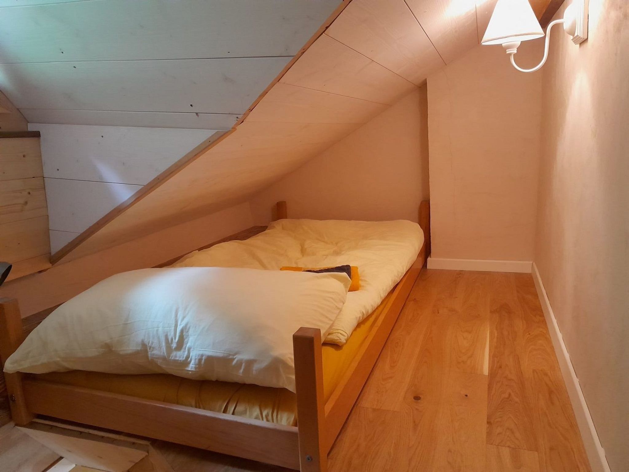 A small, single-bed room with a sloped ceiling, wooden flooring, and a wall-mounted light fixture.