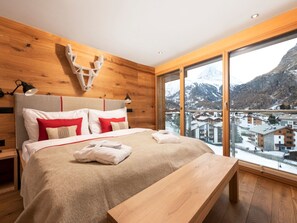 A modern bedroom with a large bed, wooden headboard, and a view of a snowy mountain town. - Chalet Nepomuk, (Zermatt), 1632.00 by Interhome (Zermatt)
