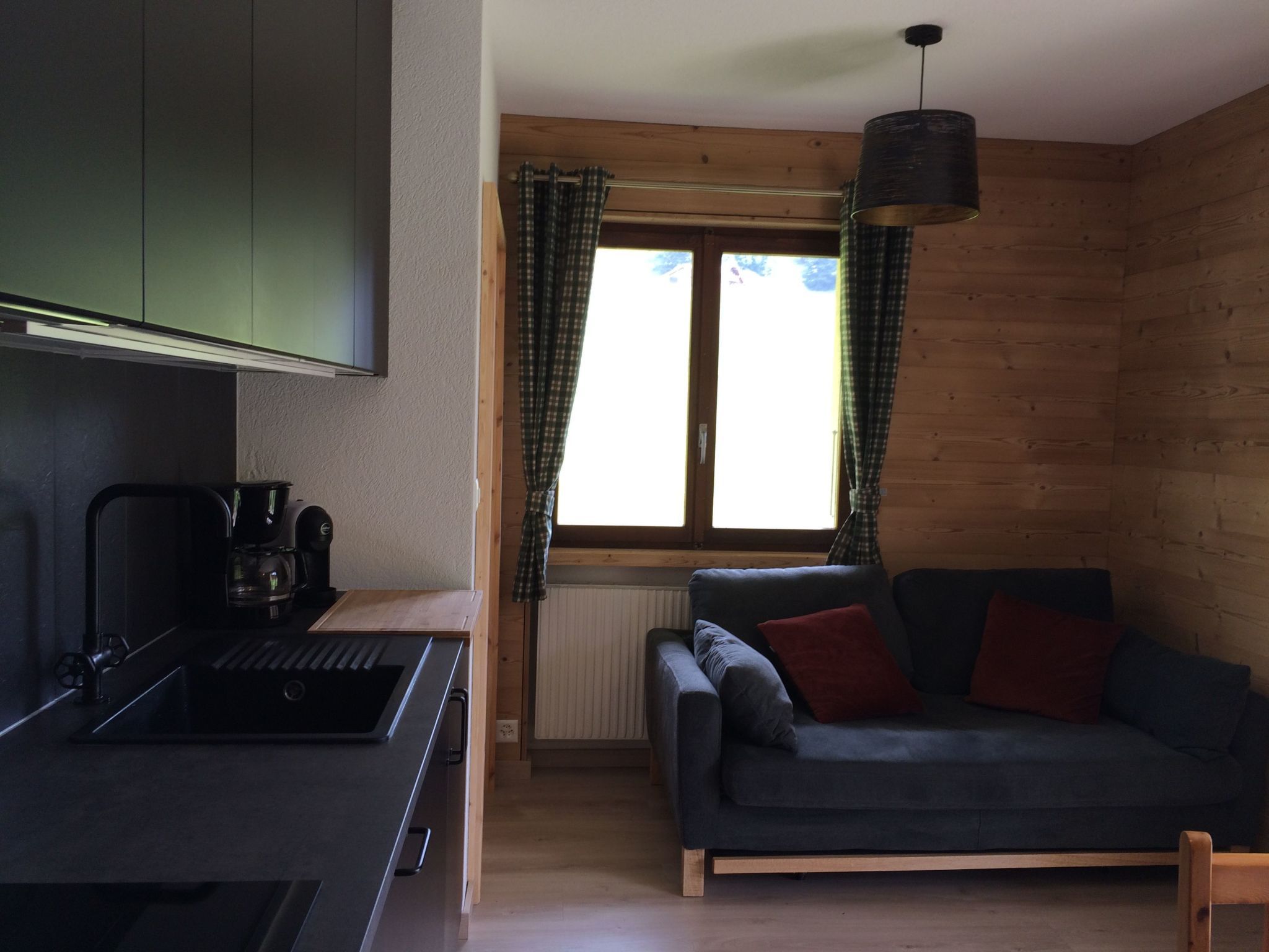 Ein kompakter Wohnbereich mit Küchenzeile, einer Couch, einem Fenster mit Vorhängen und einer Pendelleuchte.