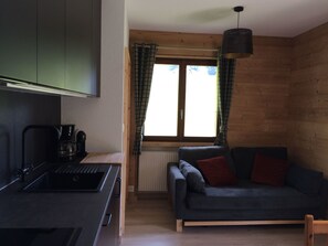 Een ruimtelijke leefruimte met een kitchenette, een bank, een raam met gordijnen en een hanglamp.