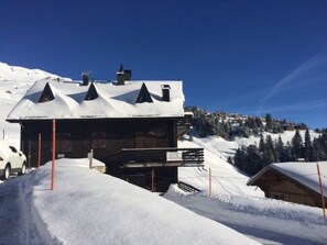 Ein schneebedecktes Gebäude mit geneigtem Dach und mehreren Schornsteinen, eingebettet in eine verschneite Landschaft unter blauem Himmel.