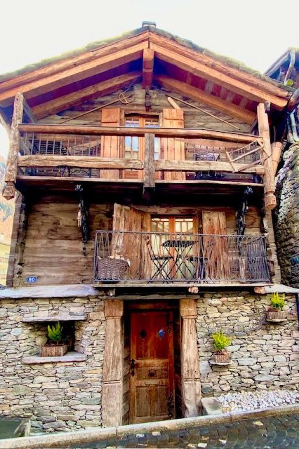 A two-story wooden house with a stone facade, a balcony with metal railings, and a red door.