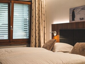 A hotel room with a bed, a window with blinds, a curtain with a pattern, a wall-mounted light, and a painting of penguins. - Haus Venus, (Zermatt), by Interhome (Zermatt)