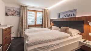A modern hotel room with a large bed, a wooden dresser, a desk, and a window with curtains. - Haus Venus, (Zermatt), by Interhome (Zermatt)
