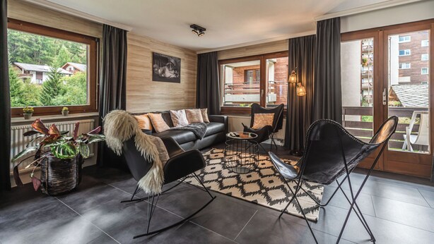 A modern living room with a black sofa, a rocking chair, a coffee table, and a view of a balcony with a railing. - Haus Venus, (Zermatt), by Interhome (Zermatt)